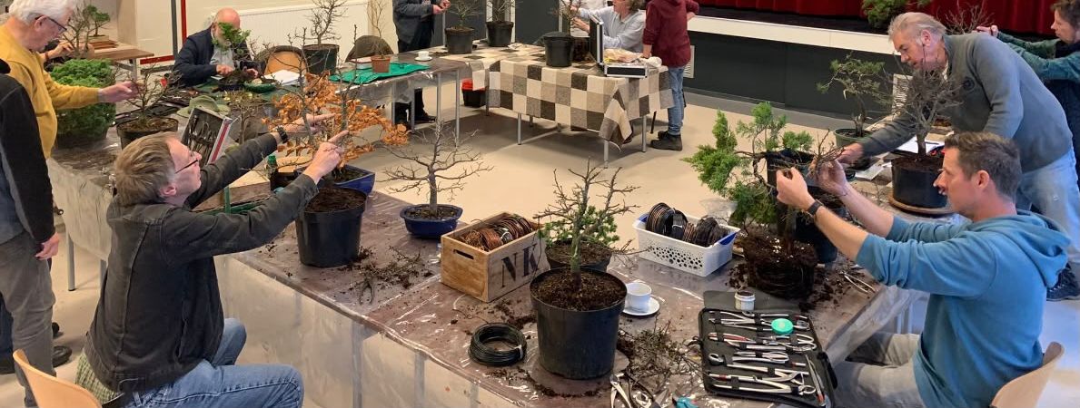 Leden werken aan hun bomen aan lange tafels in een zaal, met draad, gereedschap en koffie binnen handbereik.